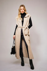 full view of long beige knit vest with waist tie over a black turtleneck and pants, styled in modern western fashion